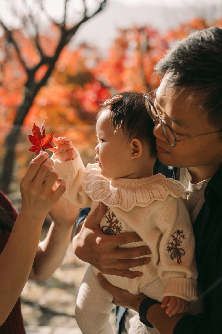 Parent holding a baby who is reaching toward a red autumn leaf during a Seoul family photoshoot