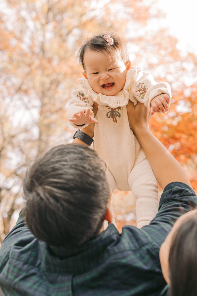 Parent lifting a laughing baby beneath colorful autumn leaves in Seoul