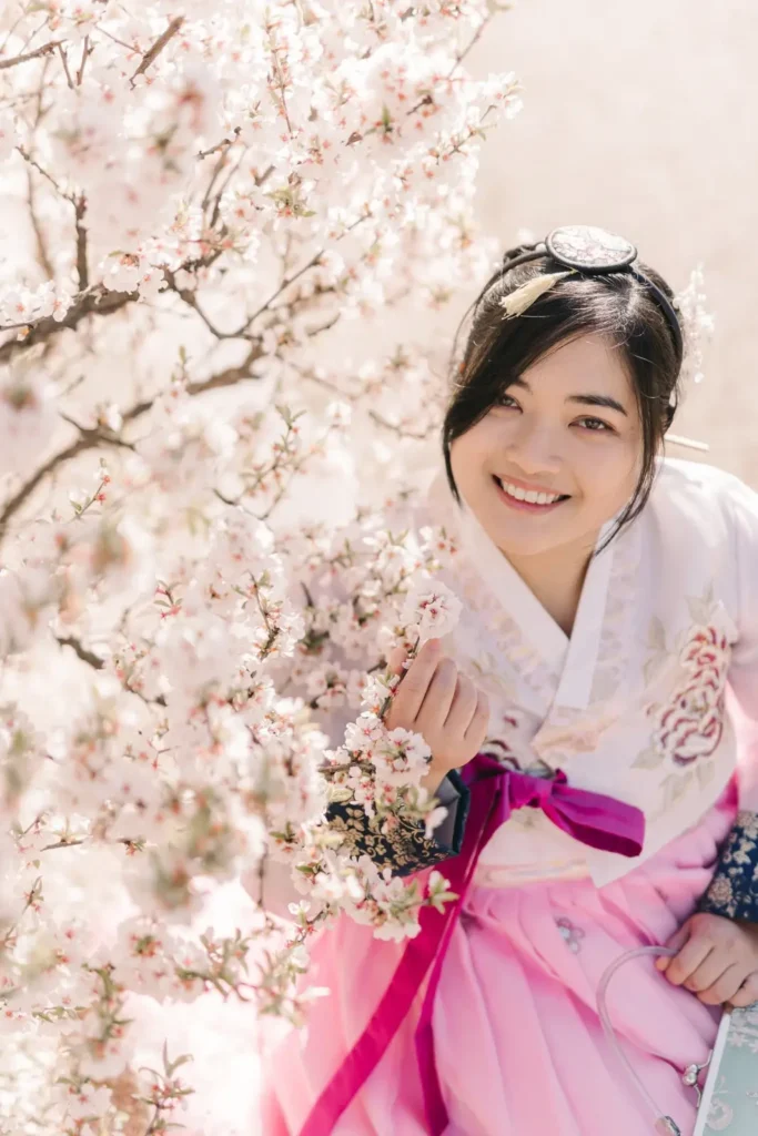 Smiling woman in a pink Korean hanbok posing beside cherry blossoms in Seoul