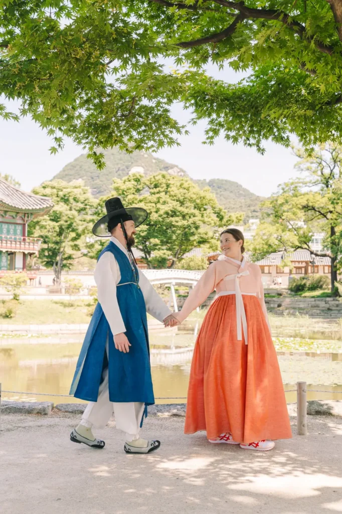 Couple holding hands in blue and orange traditional Korean hanbok beside a palace pond